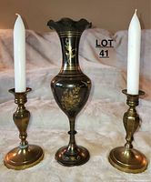 Front view of black brass vase with gold floral details flanked by two brass candlesticks holding white taper candles on a soft surface