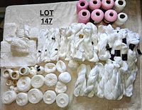 Top view of multiple yarn skeins and balls, mostly white with some pink, including labeled skeins and sample crochet pieces.