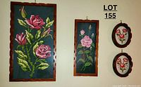 Three hanging framed floral textile artworks on wall showing one large rectangular cross stitch, one smaller rectangular cross stitch, and two oval embroidered floral pieces in wooden frames