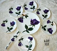 Full view of dessert set including large platter, six dessert plates, and serving spatula on a white fabric background. Each piece decorated with purple roses and green leaves.
