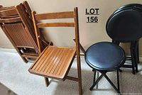 Photo showing wooden folding chair open and several folded wooden chairs stacked upright, along with three black folding stools stacked behind