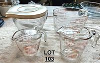 Photo showing three glass measuring cups (Pyrex) and one larger glass bowl with white lid (Anchor Hocking) all on a fabric surface, used condition.