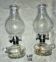 Pair of clear glass oil lamps showing front view, both lamps with round bases, loop handles, clear chimneys and metal burner assemblies with wicks.