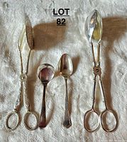 Photo showing the entire lot of silverware including salad tongs, cake server, sterling silver spoon, and silver plated spoon laid out on a textured white fabric.