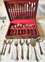 Photo showing wooden box open with 12 dinner knives in upper compartment, assorted forks and spoons in lower compartment against red felt lining, with additional serving utensils laid out in front on white fabric.