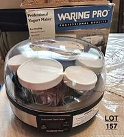 Yogurt maker unit with jars inside and clear plastic cover on top, showing brand and model details on box in background.