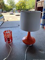 Image showing two orange lamps on a concrete surface by a large window: the smaller lamp with multiple hanging rectangular orange pieces, and the larger lamp with a solid tulip-shaped base and white fabric shade.