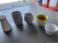 Photo showing five decorative items: two pottery face vases, one green-rimmed pottery vase with mosaic tiles, one white ceramic popcorn bowl, and one cream jug with metal handle.