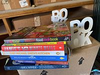 Stack of 7 cookbooks with different colors and titles on a wooden surface with two white block letter bookends spelling 'BOOK' behind them.
