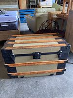 Front view of a vintage antique steamer trunk showing beige canvas, natural wood slats, and black metal reinforcements with leather handles.