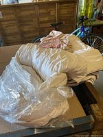 Two queen-size cream/white comforters with plastic wrapping on one, placed on a cardboard surface with some folded bedding items on top. Background shows bicycle and wooden furniture.