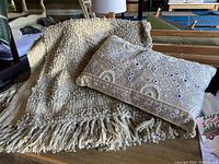 Photo of a cream textured knit blanket with fringe and a rectangular beige pillow adorned with beads, sequins, and an intricate decorative pattern.