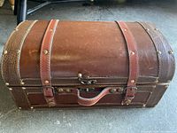 Closed view of brown mini chest made of leather featuring metal studs, leather straps, and central handle.