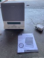 White Homedics Deep Sleep Therapy Machine with power adapter and manual placed on a surface, showing the front speaker grille, LCD screen, and control buttons.