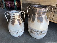 Photo showing two Egyptian style metal jugs with handles, distressed white and dark metal finish, decorative banding around lower midsection.