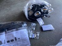 Photo of heavy duty black solar string lights coiled with clear bulb covers, small black solar panel, remote control, mounting hardware in plastic packaging, and instruction manual on concrete floor.