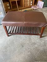 Brown leather bench with wooden slatted bottom shelf on concrete floor, background showing other furniture and boxes.