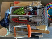 Overview of metal wire baskets with various small kitchen utensils including peelers, slicers, garlic press, skewers, and measuring cups.