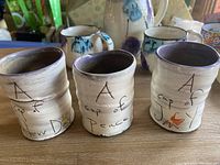 Close-up of three pottery mugs with inscriptions 'A cup of New Day', 'A cup of Peace', and 'A cup of Joy' featuring a rustic finish and star/sun designs.