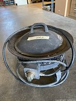 Propane fire pit placed on concrete floor, showing lid on top, attached hose coiled around, and control knob visible.