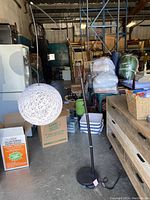 Full view of the tall floor lamp featuring a white spherical woven dome shade, tall adjustable black metal stem, and round black base in a warehouse setting.