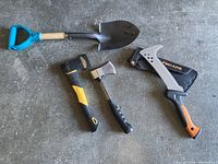 One mini shovel with blue handle, two hatchets, and a Fiskars heavy duty limb cutter with protective sheath on concrete floor.