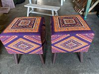 Two square fabric foot stools with African geometric pattern upholstery and dark wood legs on concrete floor with light and shadow.