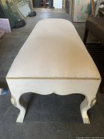 Photo showing front view of vintage cream fabric bench with curved seat, cream painted cabriole legs wrapped in leather with gold accents and floral carvings.
