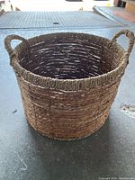 Round woven whisker basket with large side handles, viewed on a concrete floor.