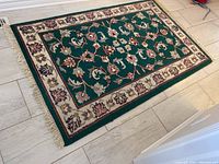 Full view of the rectangular patterned area rug laid on floor showing green central field with floral pattern and fringed edges.