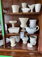 Photo of the lower two shelves of a wooden display cabinet showing a variety of white pottery items including vases, a sauce boat, planters, and decorative bowls in different shapes and textures.