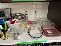 Photo showing the full baking lot including measuring spoons, Pyrex measuring cups, glass loaf pan, metal colander, mixing bowls, salt and pepper shakers, a glass oil/vinegar dispenser, vintage recipe cards, and various glass baking dishes.