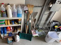 Full display of cleaning, yard, and car maintenance items arranged near garage wall including brooms, rake, shoe rack, and plastic storage bins.