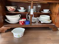 Photo shows wooden cabinet with multiple white serving bowls, plates, mugs, and some glassware partially stacked or nested. Large white oval serving bowls, white bowl with red lobster design, and a milk glass bowl with holly pattern below shelf.
