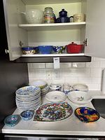 Photo showing shelves with assorted pottery bowls, mugs, teapot and a colander, along with various plates and bowls on the counter below.