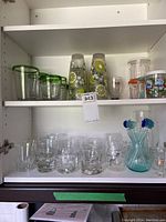 Front view of glassware on shelves including glasses with green rims, lemon motif glasses, shot glasses, plastic glasses with lids, and glass stir sticks in a pitcher.