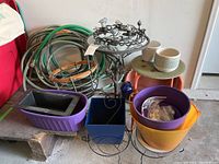 Overview photo showing metal and glass plant stand with accompanying planters in various materials and colors, metal bird candle holder, watering hoses coiled behind, and an orange metal stool.