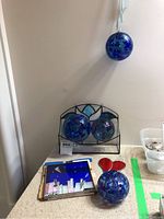 Photo showing three blue-purple hanging glass balls; two placed inside a mounted metal display box and one on a table near a red glass heart and a square stained glass art piece.