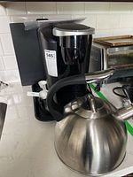Photo showing a black Keurig coffee maker with silver handle on top, pod holder with coffee pods and a white coffee measuring scoop in front, alongside a stainless steel whistling kettle with black handle.