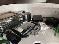 Photo showing all five kitchen appliances arranged on countertop: George Foreman grill, GE panini press, Black & Decker toaster, Cuisinart toaster oven, and white handheld mixer.