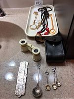 Overview of the lot showing the Alpaca silver comb case, USSR opera glasses with carrying case, decorative spoons, and various necklaces.