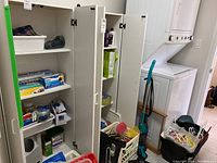 Two white tall cabinets filled with cleaning supplies, laundry hampers, and household items, with visible stacked washer and dryer nearby.