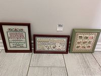 Photo of three framed needlepoint/cross stitch samplers against a wall on tile floor.