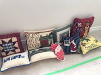 Photo of eight small decorative pillows arranged on floor showing various designs including rug hooked, quilted patchwork, hand-stitched, and embroidered text.