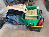 Wide assortment of reusable bags, a green plastic shopping basket, a clear plastic storage tote, and a black plastic storage tote with some fabric bags inside all on floor.