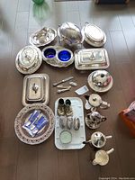 Full overview of all silver plated items laid out on floor showing serving dishes with lids, tea pots, salt & pepper shakers, table lighters, silver spoons in packaging, and other utensils.
