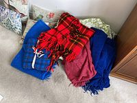 Group photo showing six assorted throw pillows with different patterns and colors, and four throw blankets piled including red plaid, blue plaid, solid blue, and solid reddish colors.