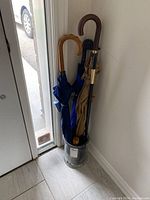 Five umbrellas standing inside a tall, clear glass umbrella stand by the door.