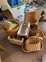Photo showing a cluster of various wicker baskets in different shapes and sizes, placed on the floor around a glass table and some plastic storage containers.