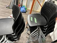 Stack of 10 black stationary office chairs showing plastic seats and perforated backs, on metal legs, stacked in a room with wood floor.
